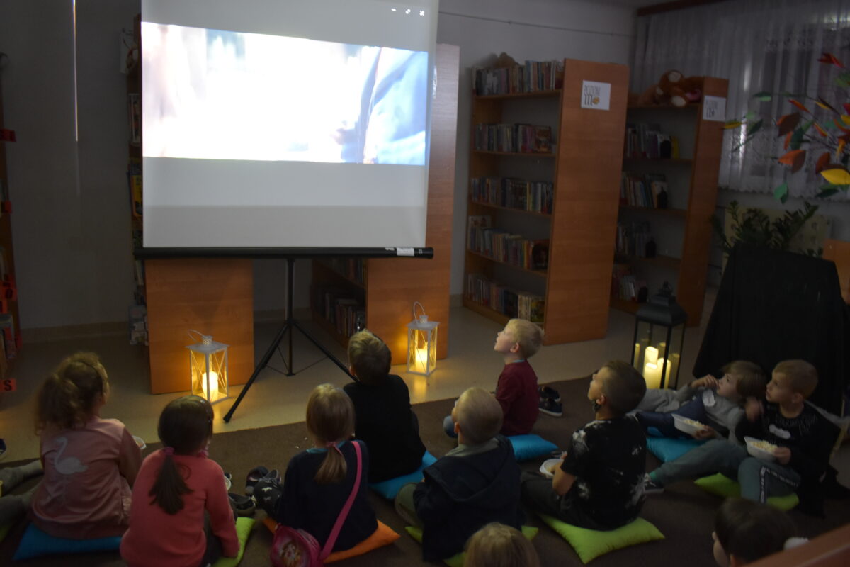 Grupa dzieci siedzi na wykładzinie, oglądają film, w tle regały z książkami i świecące lampiony