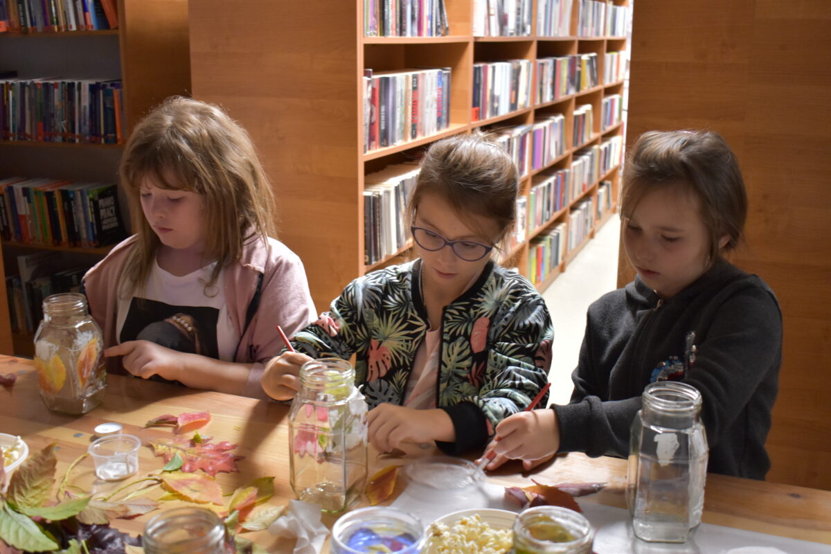Trzy dziewczynki malują słoiki i przyklejają na nich kolorowe liście, za nimi stoją regały z książkami.