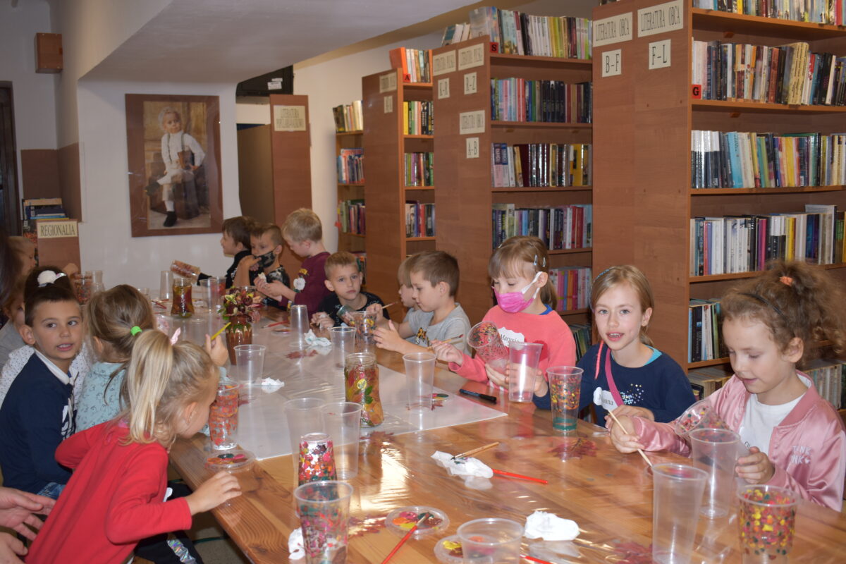 Przy długim stole siedzą dzieci, które malują farbami plastikowe kubeczki. W tle regały z książkami