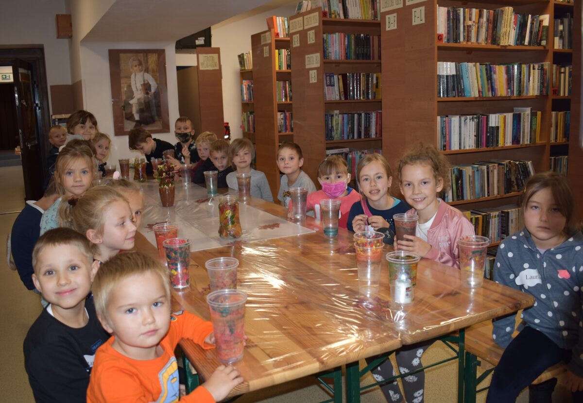 Zadowolona gromadka dzieci siedzi przy stole, w ręku trzymają plastikowe kubeczki pomalowane w barwy jesieni. W tle półki pełne książek.