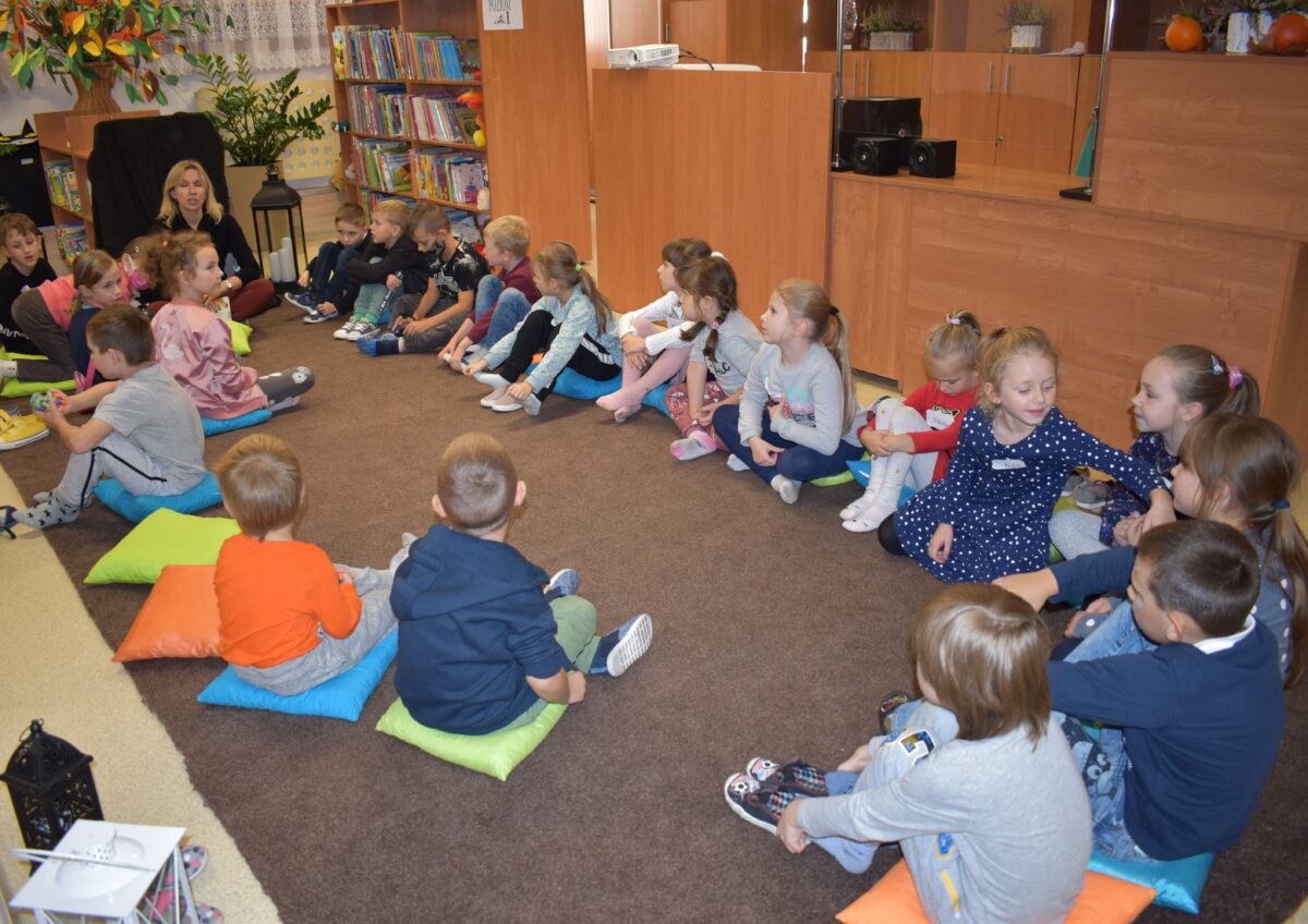 W sali bibliotecznej na kolorowych poduszkach siedzą dzieci i słuchają pani która do nich mówi. W tle regały i półki z książkami oraz kwiaty.