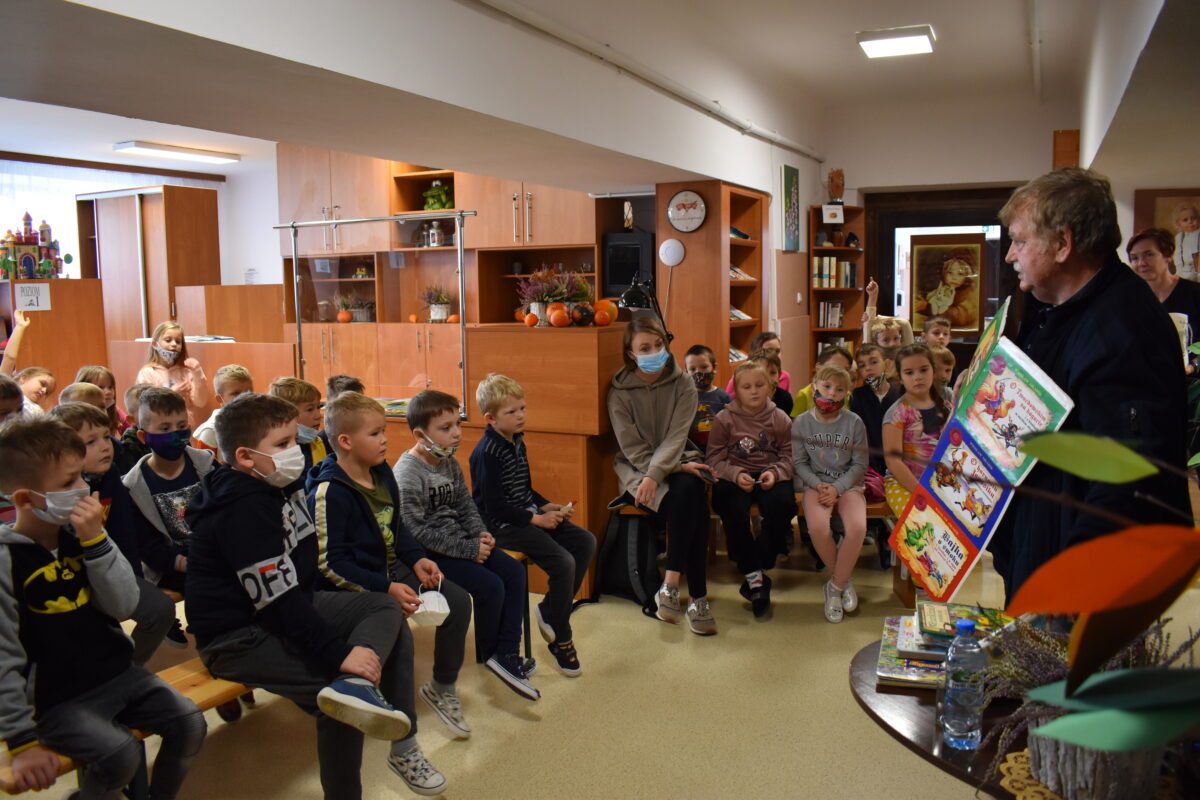 Grupa dzieci stojących i siedzących przy nich stoi mężczyzna, w oddali widać regały z książkami, czarną kotarę, i kolorowe liście