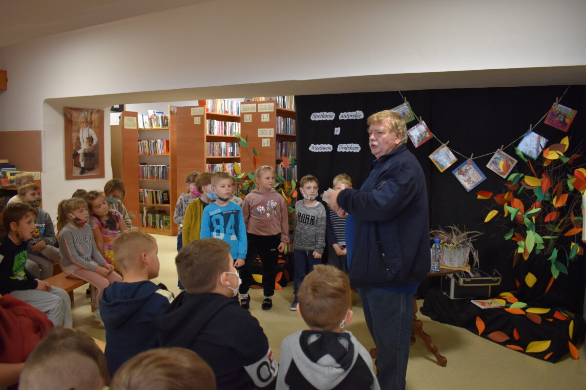 W bibliotece dziewczynki i chłopcy siedzą na podłodze, kilkoro z nich stoi obok nich autor książek wszyscy z nich trzymają w ręku dyplomy. Dalej widać regały z książkami, czarną kotarę na której wiszą książki a obok w wazonie stoją kolorowe liście