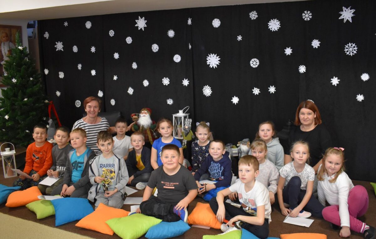 Na dywanie siedzą dzieci i pani która pokazuje książkę. Obok niej stoją kolorowe krasnale, białe lampiony, sto