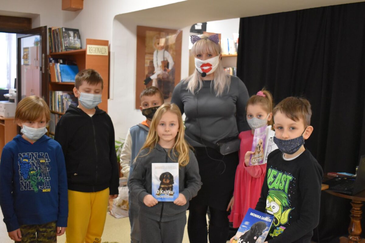 Grupka dzieci z panią , troje z nich trzyma książki. W tle regał z książkami
