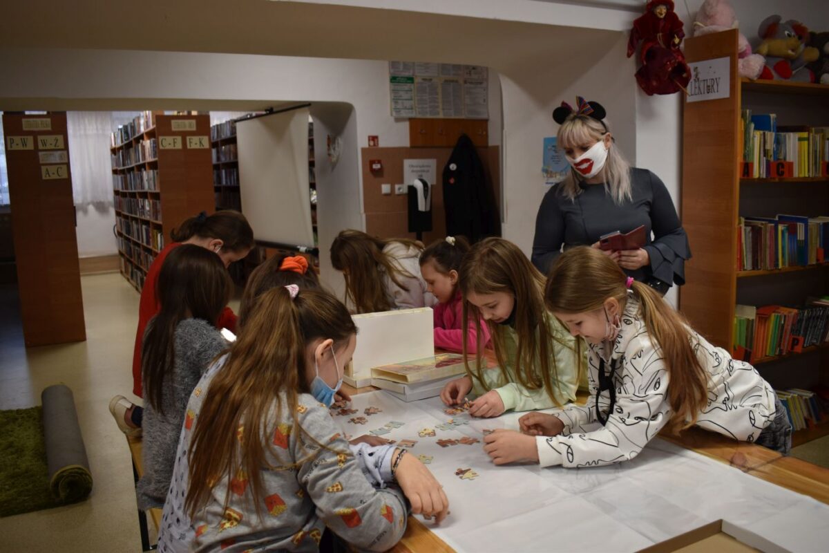 Dziewczynki pochylają się nad stołem na którym układają puzzle. Przygląda im się kobieta z maską na twarzy i opasce w kształcie uszek myszki. W tle regały z książkami