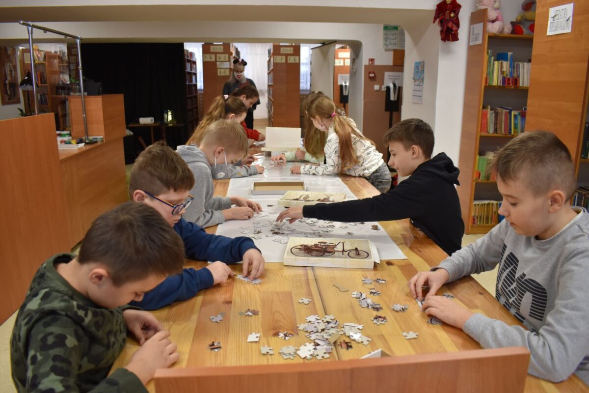 W sali bibliotecznej grupa dzieci układa puzzle w tle książki stojące na półkach.