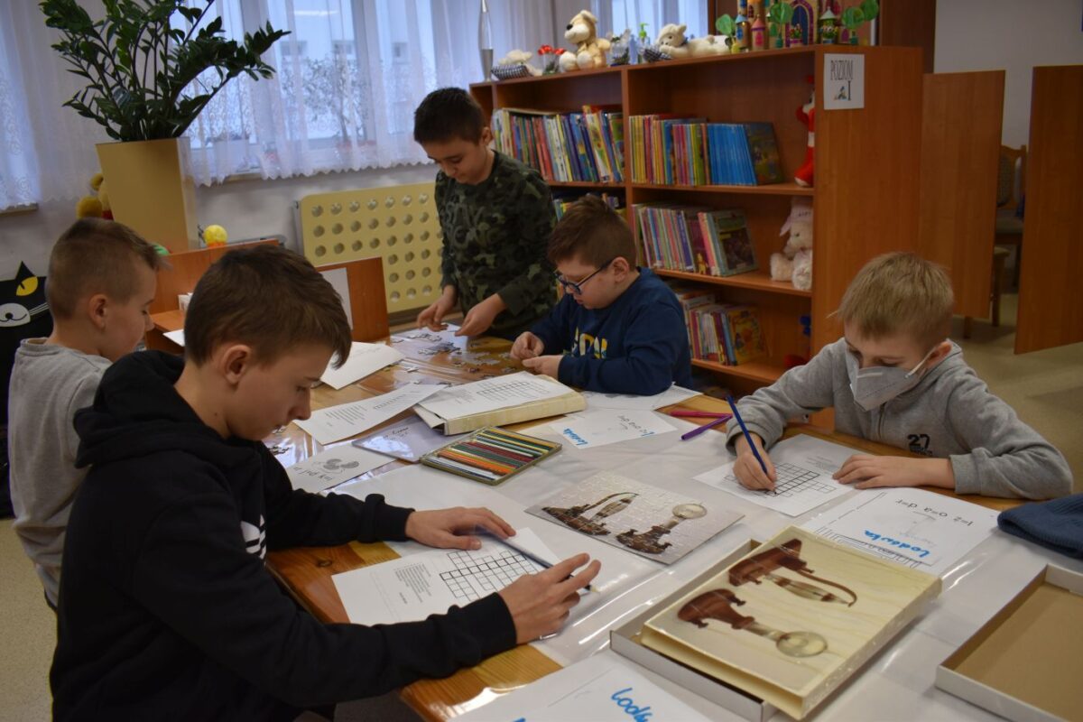 Chłopcy siedzą przy stole i rozwiązują krzyżówki. Na stole leżą kartki, kredki, rysunki, w tle półki z książkami