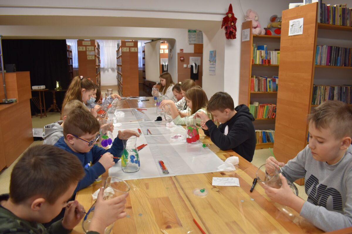 W bibliotece przy długim stole siedzą dzieci i malują na szklanych karafkach kolorowe owoce. W tle wiele regałów z książkami.