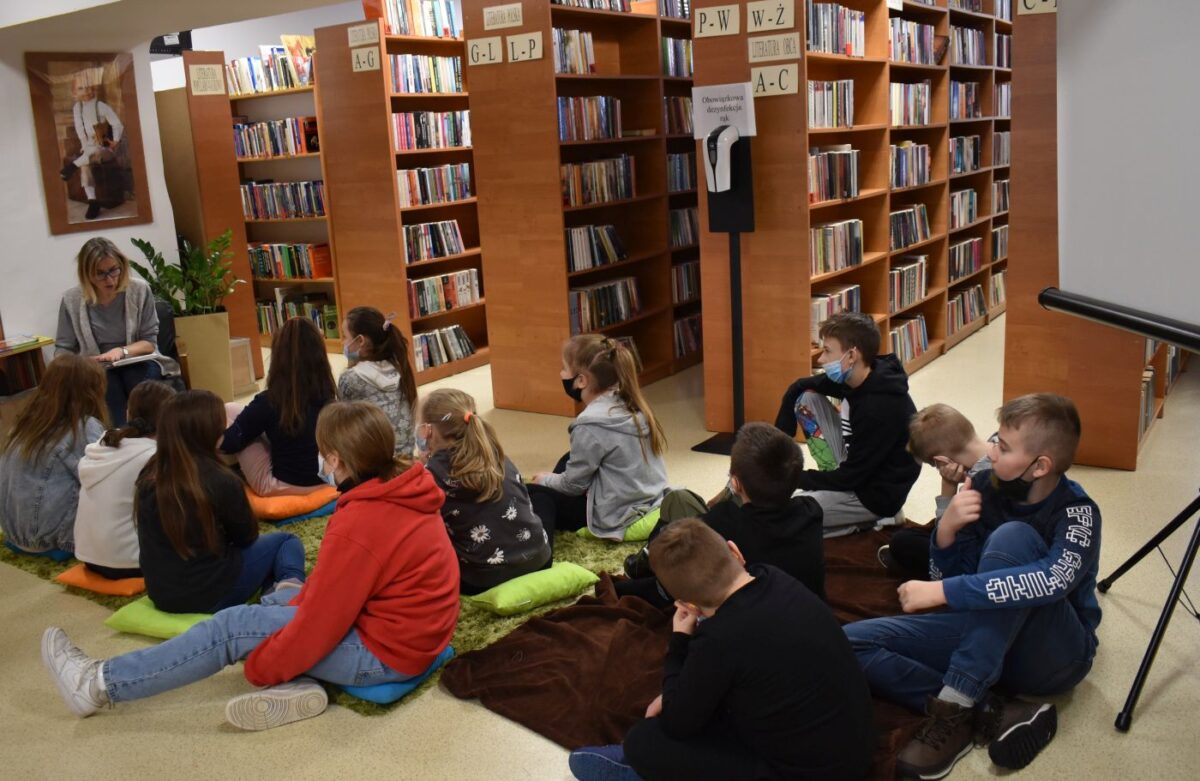 Grupa dzieci przykleja do talerzyków papierowe serwetki przed nimi stoją białe świece z motywami kwiatowymi. W tle regały z książkami