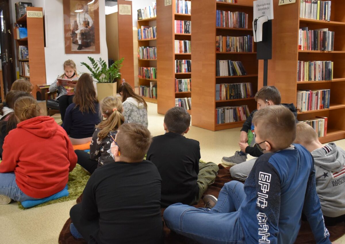 dzieci siedzą na zielonym dywanie i słuchają koleżanki która czyta książkę obok niej zielony kwiatek w tle regały z książkami
