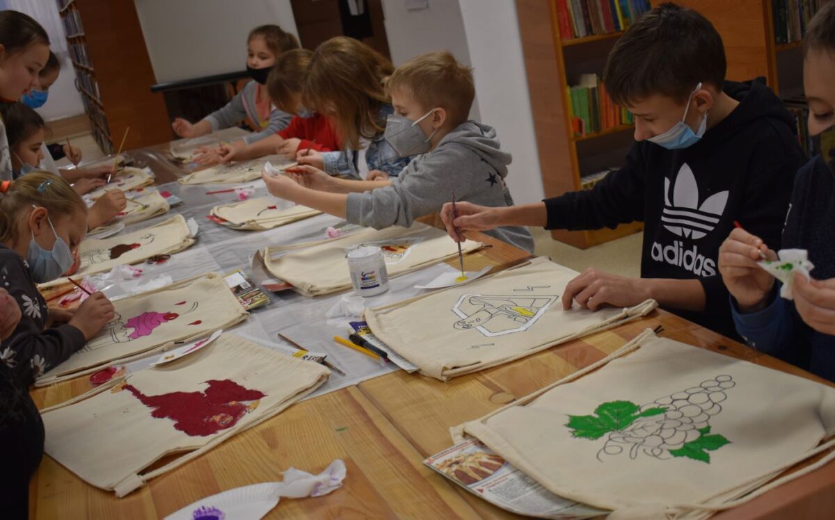Przy stole grupka dzieci maluje na jasnych workach kolorowe obrazki