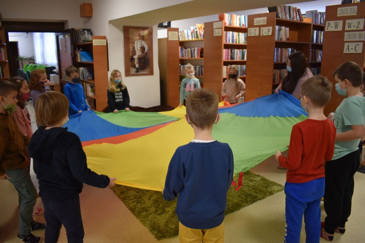 Grupka dzieci trzyma kolorową chustę w tle półki z książkami