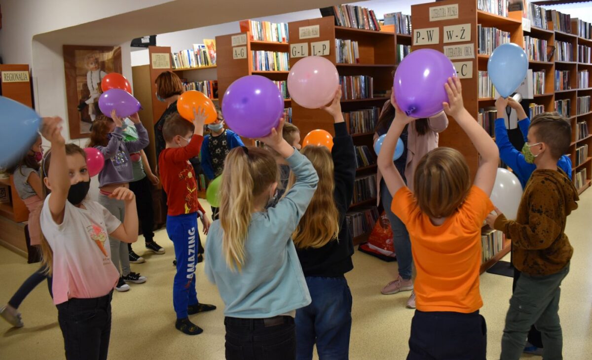Dzieci z uniesionymi rękoma w których trzymają kolorowe balony za nimi regały pełne książek