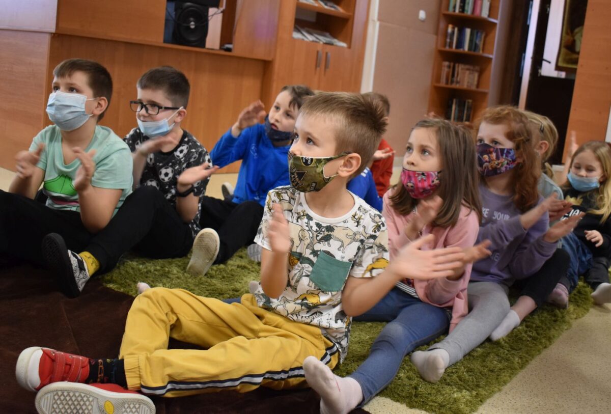 Dzieci siedzące w bibliotece na zielonym dywanie biją brawo.