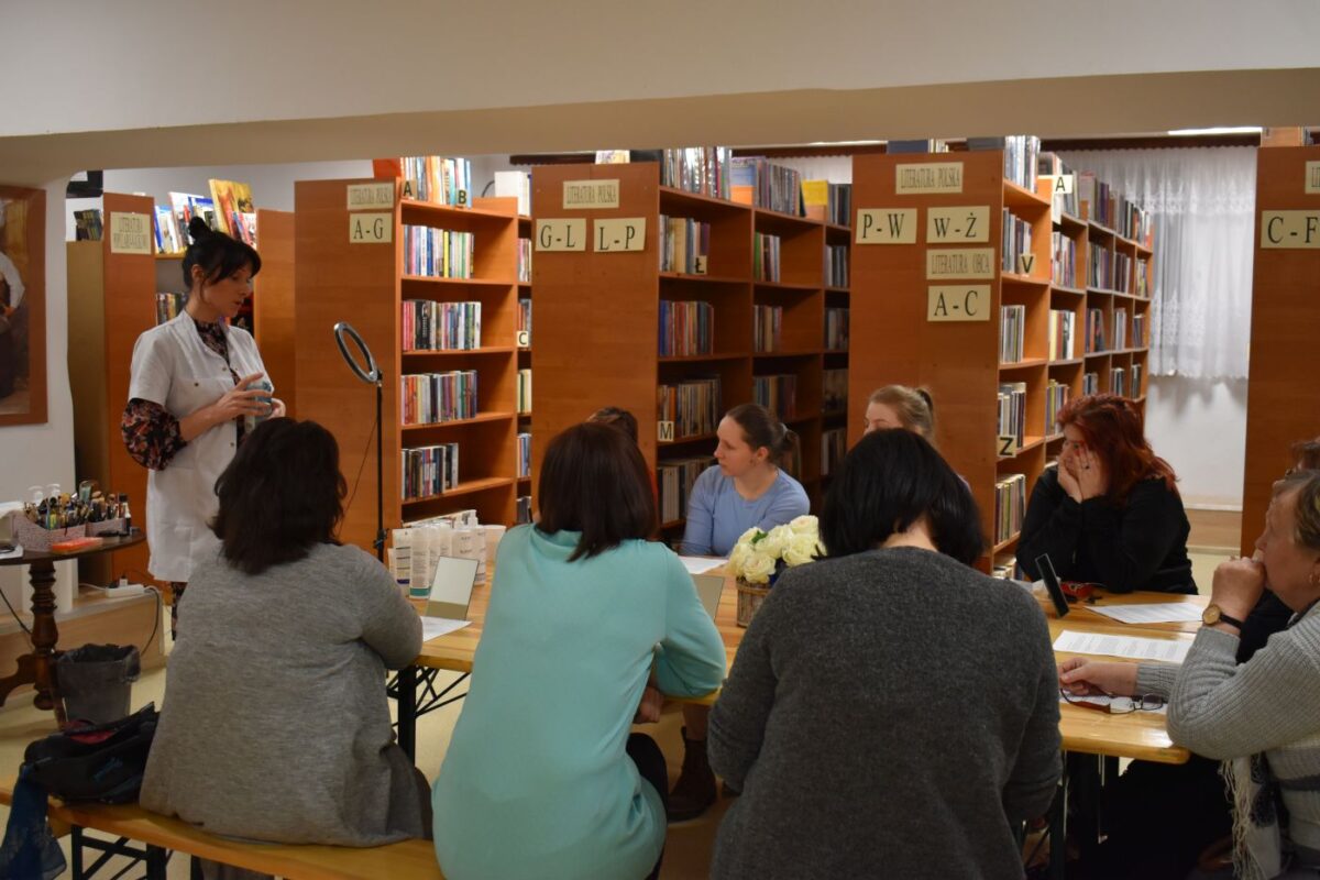 Przy drewnianym stole na którym stoją lusterka, białe kwiaty, kosmetyki siedzą kobiety i słuchają pani w białym fartuchu. W tle stoją regały.