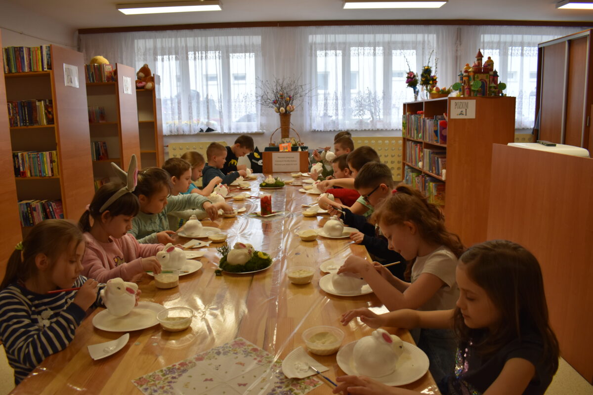 Dzieci przy drewnianym stole układają na mchu stroiki świąteczne z białymi królikami. W tle regały z książkami.