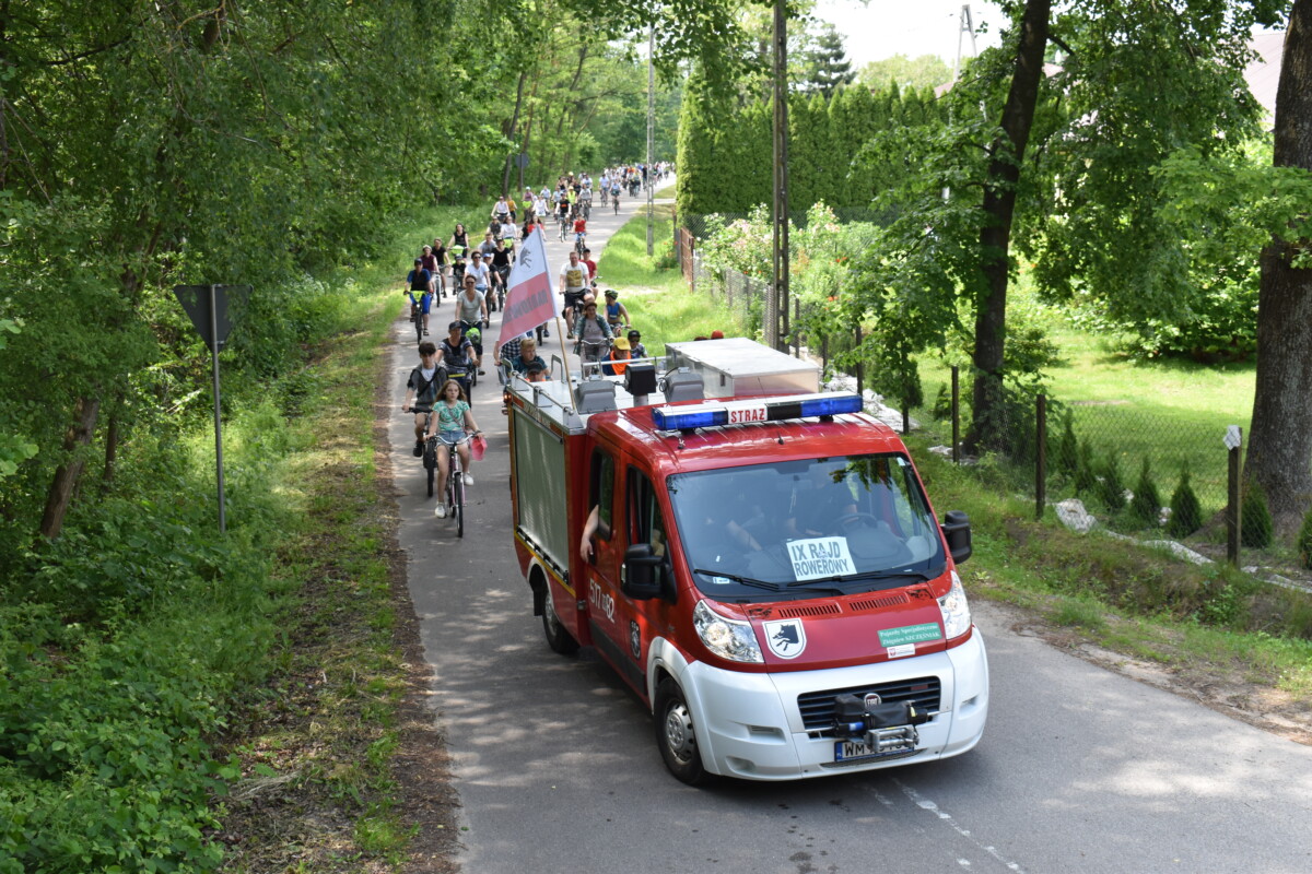 Na pierwszym planie czerwony samochód strażacki za nim peleton rowerów. Po obu stronach ulicy zielone drzewa i trawniki