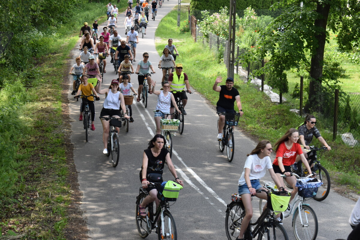 Drogą jedzie wiele uśmiechniętych osób na rowerach, niektórzy machają, po obu stronach jest zielono
