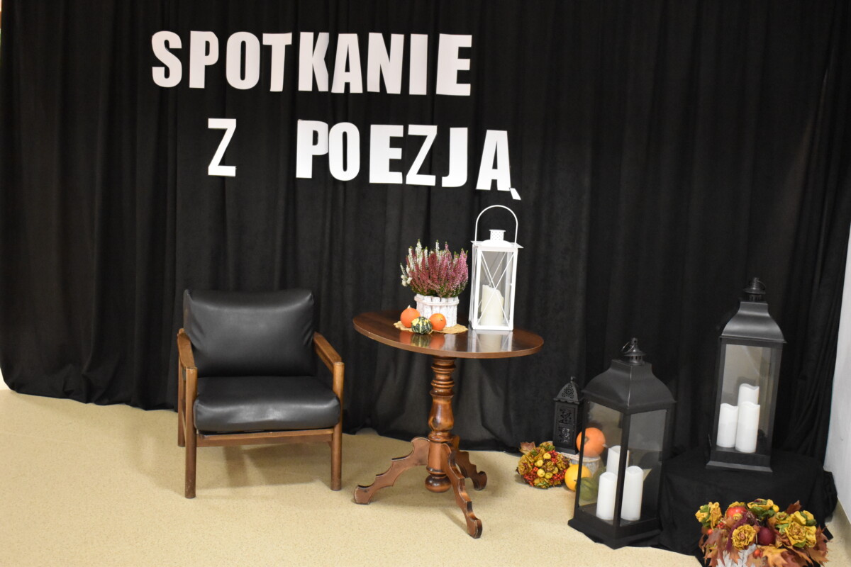 Czarna kotara na niej biały napis Spotkanie z poezją, przed kotarą leżą lampiony, dynie, fotel, stolik na którym stoi wrzos, tykwy i lampion