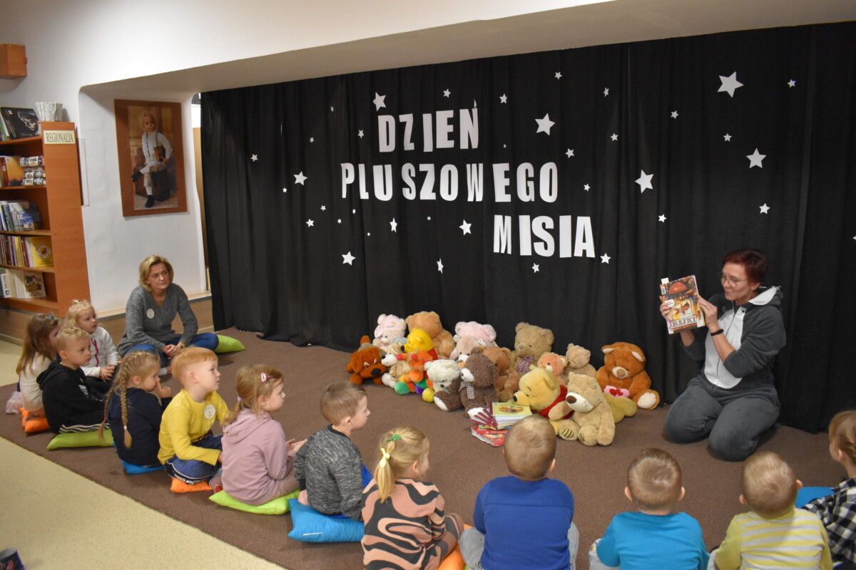 Grupa przedszkolaków i dwie panie pozują do zdjęcia. Dzieci w rękach trzymają kolorowe misie. W tle czarna kotara z białym napisem i gwiazdkami, przed nimi leżą kolorowe poduszki
