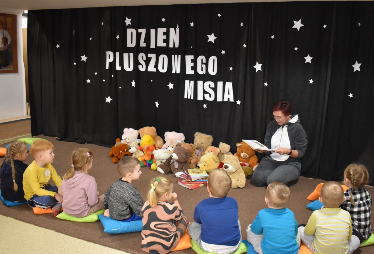 Grupa przedszkolaków i dwie panie pozują do zdjęcia. Dzieci w rękach trzymają kolorowe misie. W tle czarna kotara z białym napisem i gwiazdkami, przed nimi leżą kolorowe poduszki