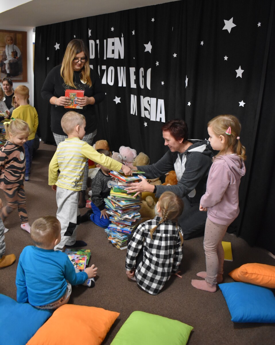 Przedszkolaki siedzą na kolorowych poduszkach i oglądają książki przed nimi leży kilkanaście misiów pluszowych w tle czarna kotara z białym napisem