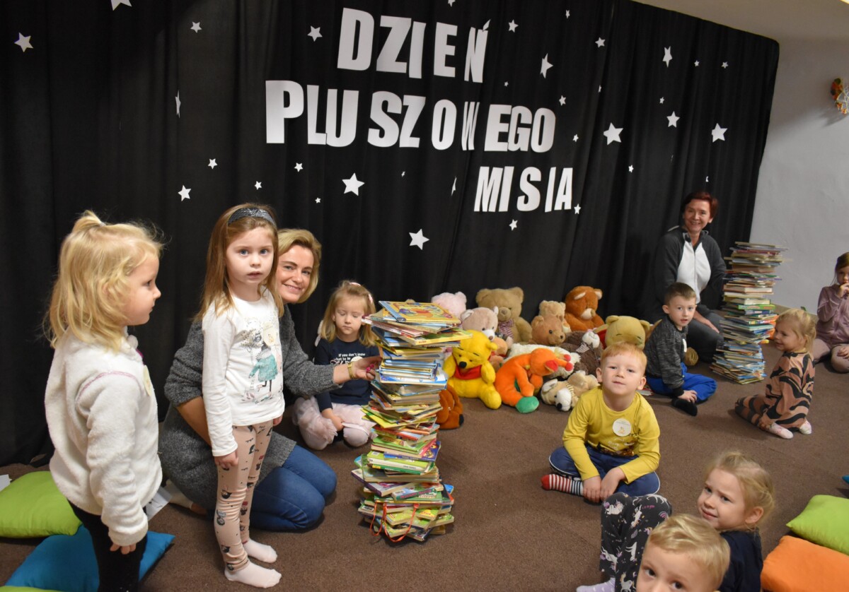 Przedszkolaki siedzą na kolorowych poduszkach i oglądają książki przed nimi leży kilkanaście misiów pluszowych w tle czarna kotara z białym napisem