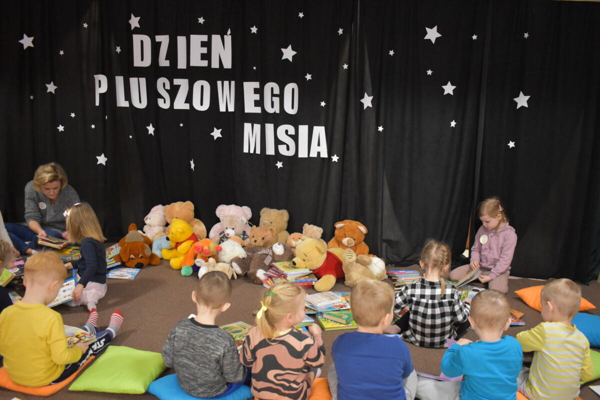 Przedszkolaki siedzą na kolorowych poduszkach i oglądają książki przed nimi leży kilkanaście misiów pluszowych w tle czarna kotara z białym napisem