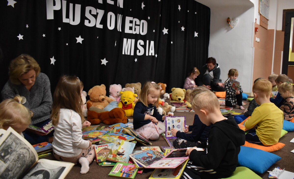 Przedszkolaki siedzą na kolorowych poduszkach i oglądają książki przed nimi leży kilkanaście misiów pluszowych w tle czarna kotara z białym napisem