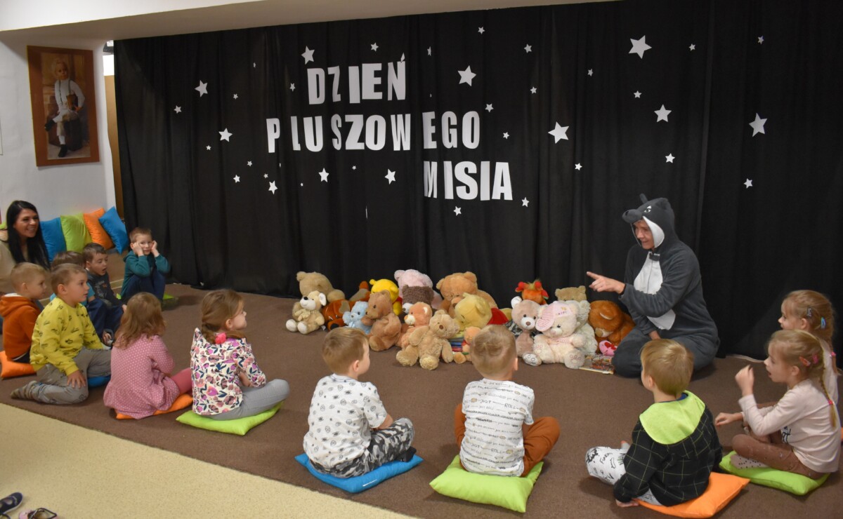 Przedszkolaki siedzą na kolorowych poduszkach i oglądają książki przed nimi leży kilkanaście misiów pluszowych w tle czarna kotara z białym napisem