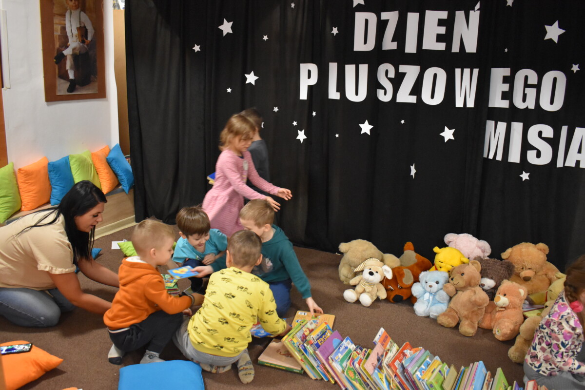 Przedszkolaki siedzą na kolorowych poduszkach i oglądają książki przed nimi leży kilkanaście misiów pluszowych w tle czarna kotara z białym napisem