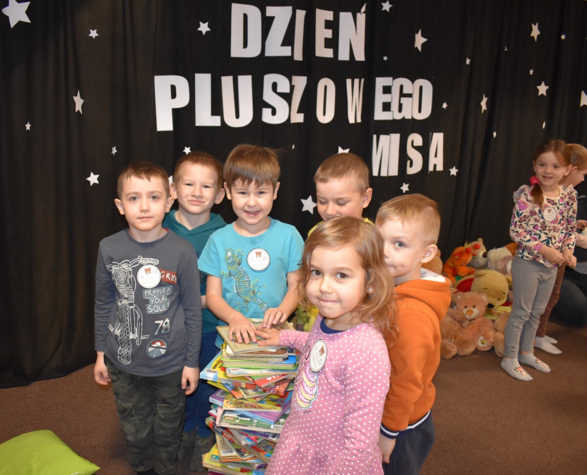 Przedszkolaki siedzą na kolorowych poduszkach i oglądają książki przed nimi leży kilkanaście misiów pluszowych w tle czarna kotara z białym napisem