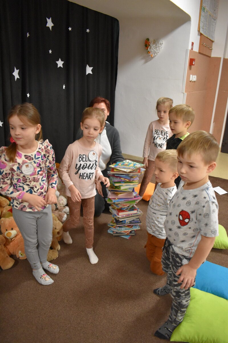 Przedszkolaki siedzą na kolorowych poduszkach i oglądają książki przed nimi leży kilkanaście misiów pluszowych w tle czarna kotara z białym napisem
