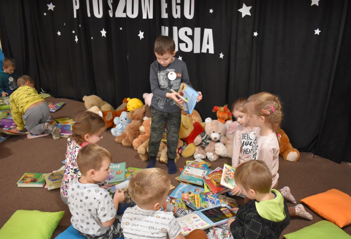 Przedszkolaki siedzą na kolorowych poduszkach i oglądają książki przed nimi leży kilkanaście misiów pluszowych w tle czarna kotara z białym napisem