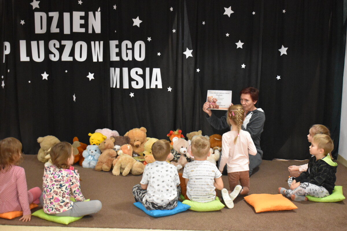 Przedszkolaki siedzą na kolorowych poduszkach i oglądają książki przed nimi leży kilkanaście misiów pluszowych w tle czarna kotara z białym napisem