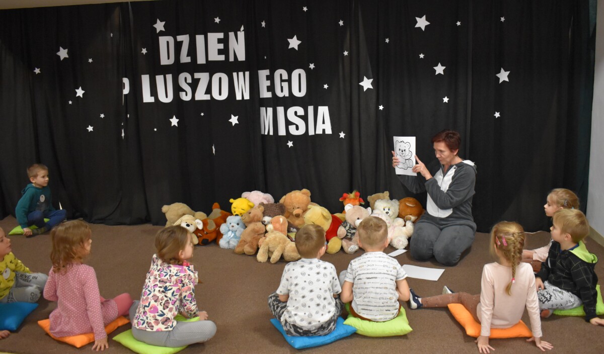 Przedszkolaki siedzą na kolorowych poduszkach i oglądają książki przed nimi leży kilkanaście misiów pluszowych w tle czarna kotara z białym napisem