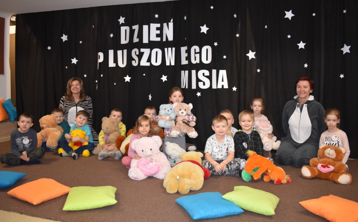 Przedszkolaki siedzą na kolorowych poduszkach i oglądają książki przed nimi leży kilkanaście misiów pluszowych w tle czarna kotara z białym napisem