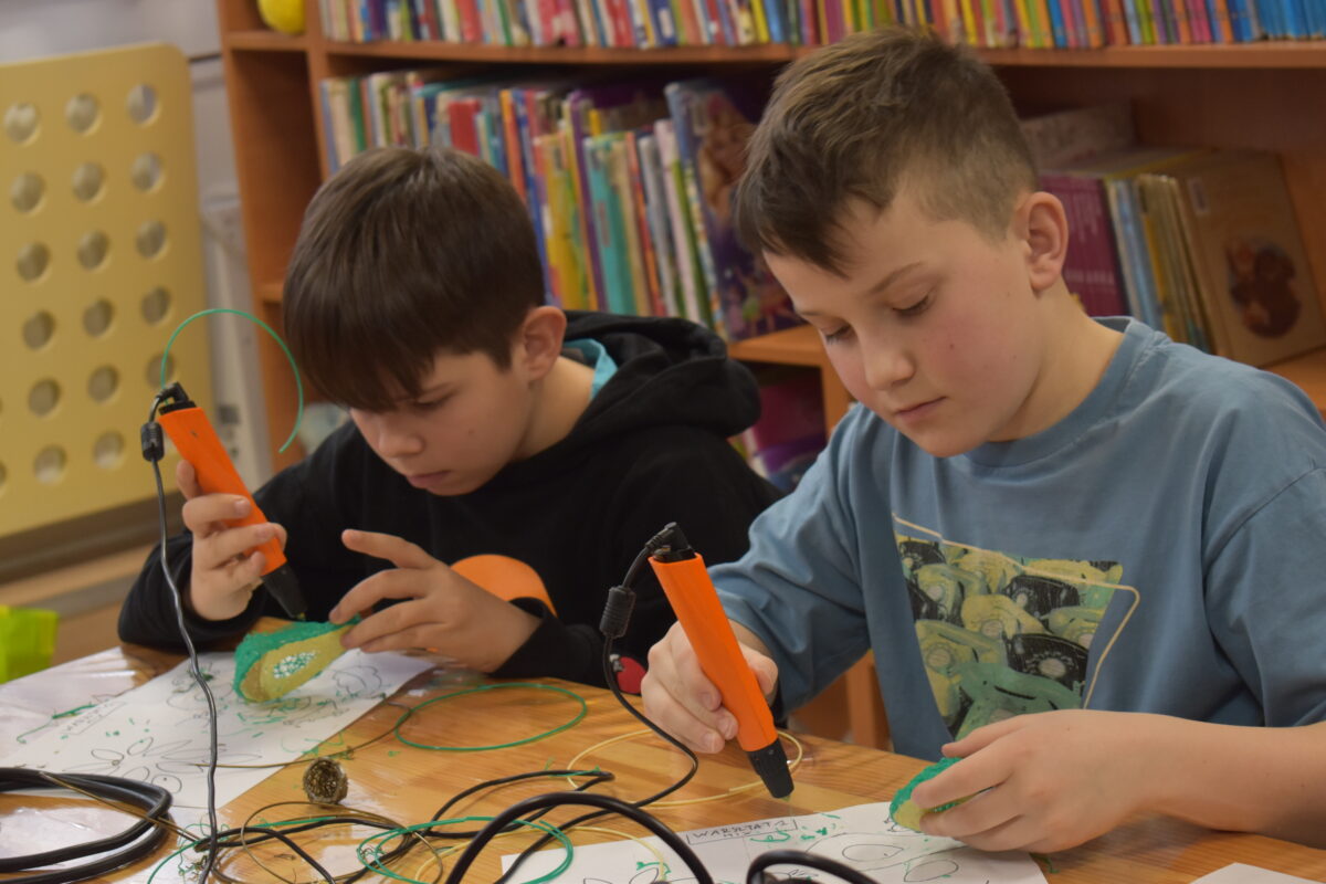 W bibliotece na zielonym dywanie siedzi kobieta i dzieci w kółku, za nimi regały z książkami.