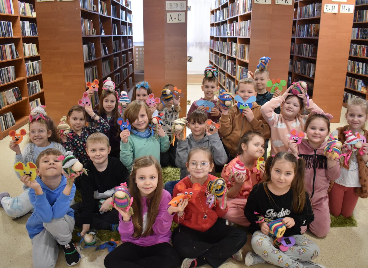 160 Uśmiechnięte dzieci pozują do zdjęcia z zrobionymi przez siebie skrzatami i magnesami. W tle biblioteczne regały z książkami.