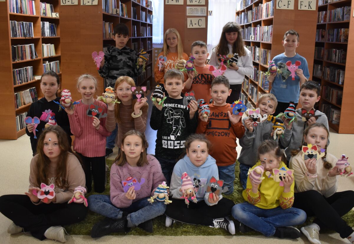 202 W pomieszczeniach biblioteki dzieci siedzą przy drewnianym stole i malują przezroczyste szklanki farbami trzymają pędzelki w dłoniach. W tle regały z książkami.