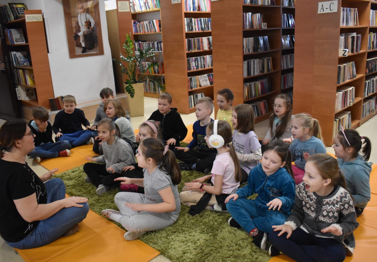 Dziewczynki i chłopcy siedzą przy stole i rysują pomarańczowymi długopisami w tle regały z książkami.