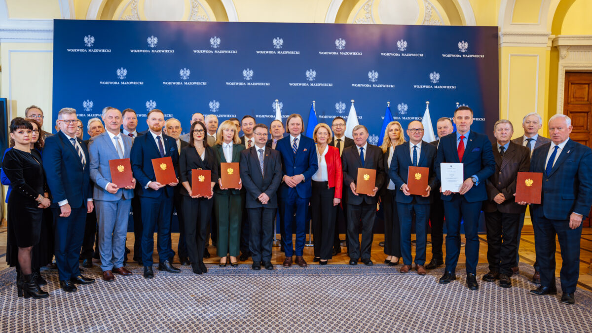 Obraz przedstawia grupę osób stojących w dwóch rzędach, ubranych elegancko, większość mężczyzn w garniturach, a kobiety w sukienkach lub eleganckich garniturach. Na pierwszym planie sześć osób trzyma certyfikaty lub podobne dokumenty w brązowych okładkach. W tle stoi drugi rząd ludzi, również z dokumentami w rękach. Wszyscy patrzą w stronę kamery z uśmiechem i wyglądają na zadowolonych. Tło stanowi wnętrze o eleganckim charakterze, prawdopodobnie sala konferencyjna lub podobne pomieszczenie. Na ścianie w tle widnieje kilka flag, co może sugerować, że jest to wydarzenie o charakterze międzynarodowym lub rządowym. Pośrodku, na ścianie, znajduje się duże logo lub herb, co może wskazywać na konkretną instytucję lub organizację. Na podłodze jest wykładzina, a całe pomieszczenie jest dobrze oświetlone. Wydarzenie to wydaje się być ważną ceremonią lub uroczystością, być może wręczeniem nagród, certyfikatów lub uznania dla grupy ludzi. Atmosfera jest formalna i celebracyjna.