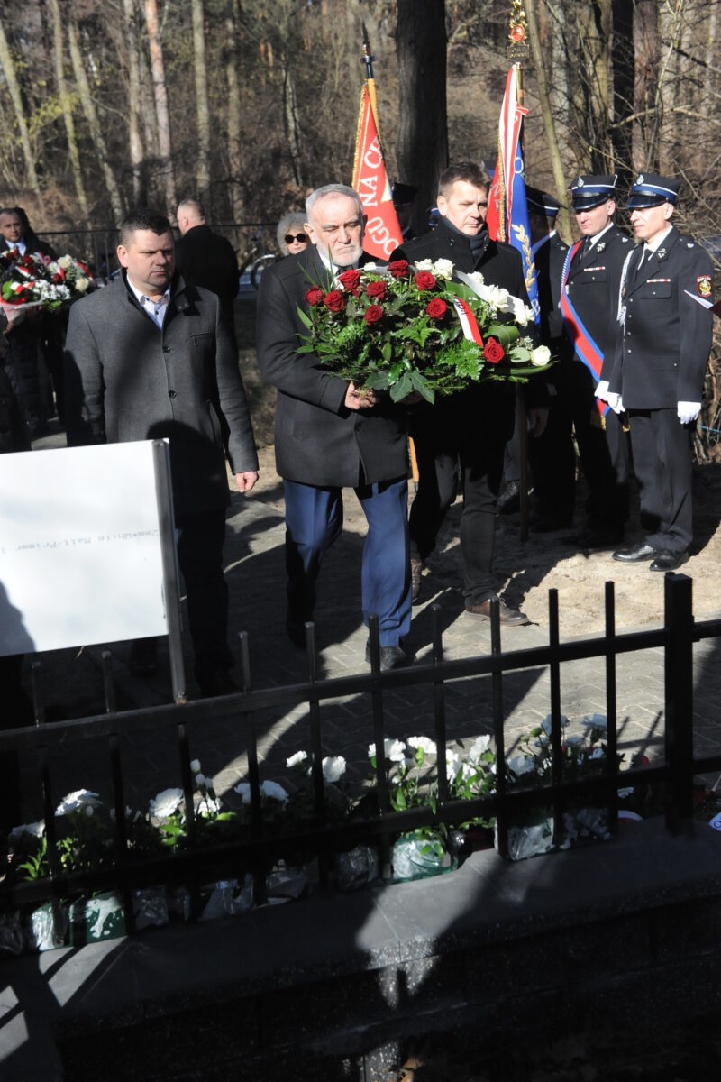 Na obrazku widać grupę osób stojących przed ogrodzeniem. Wyglądają na podejrzane lub skupione, podejrzanec, że może wykryć coś poza klastką.
