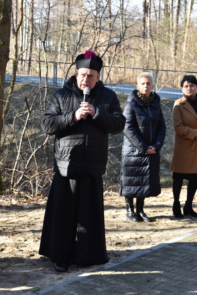 Na obrazku grupy osób, która stoi w parku. Otoczeni są zielenią drzew i różnokolorowych kwiatów, a ich twarze są całkowicie pełne radości i spokoju.