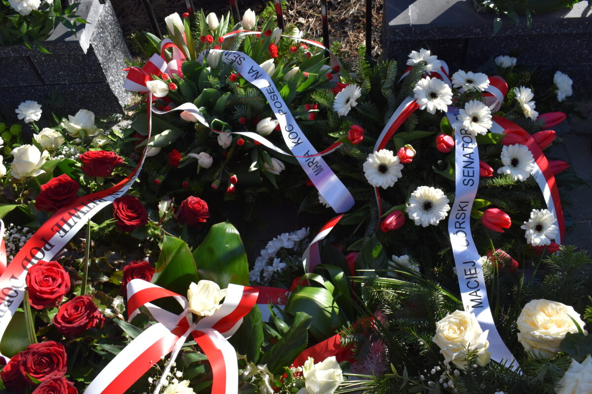 Na obrazku widocznym jest wieniec z czerwonych, białych i niebieskich kwiatów. Jest on starannie wykorzystany i zawiera różnorodne kształty i odcienie, dodając do niego głębię i różnorodność kolorów.