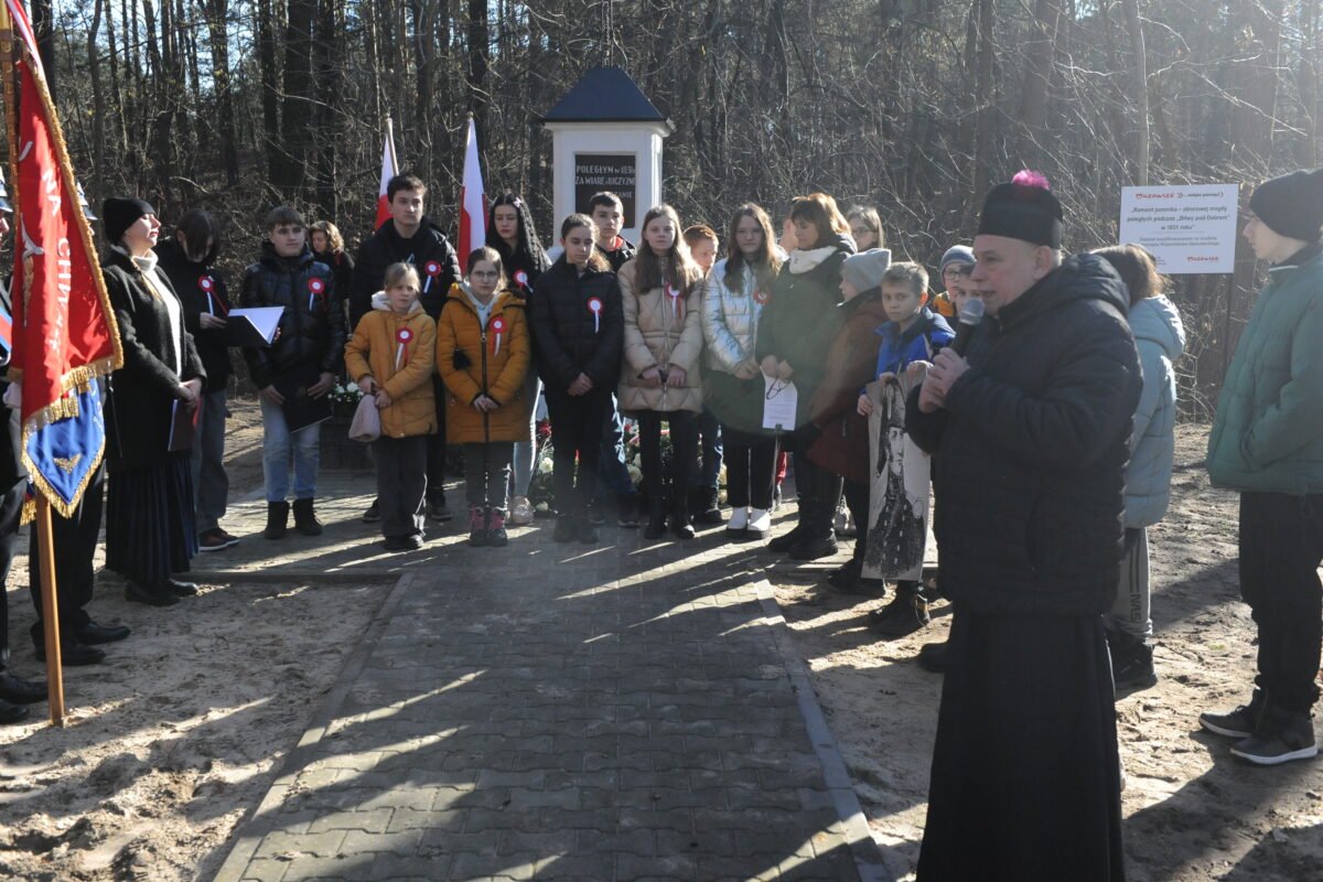 Na zdjęciu widać grupę osób, która stoi obok pomnika znajdującego się w leśnym miejscu zamieszkania. Ludzie to odrzucają i rozmawiają ze sobą, gdy pomnik wydaje się być starym i krytykowanym mchem.