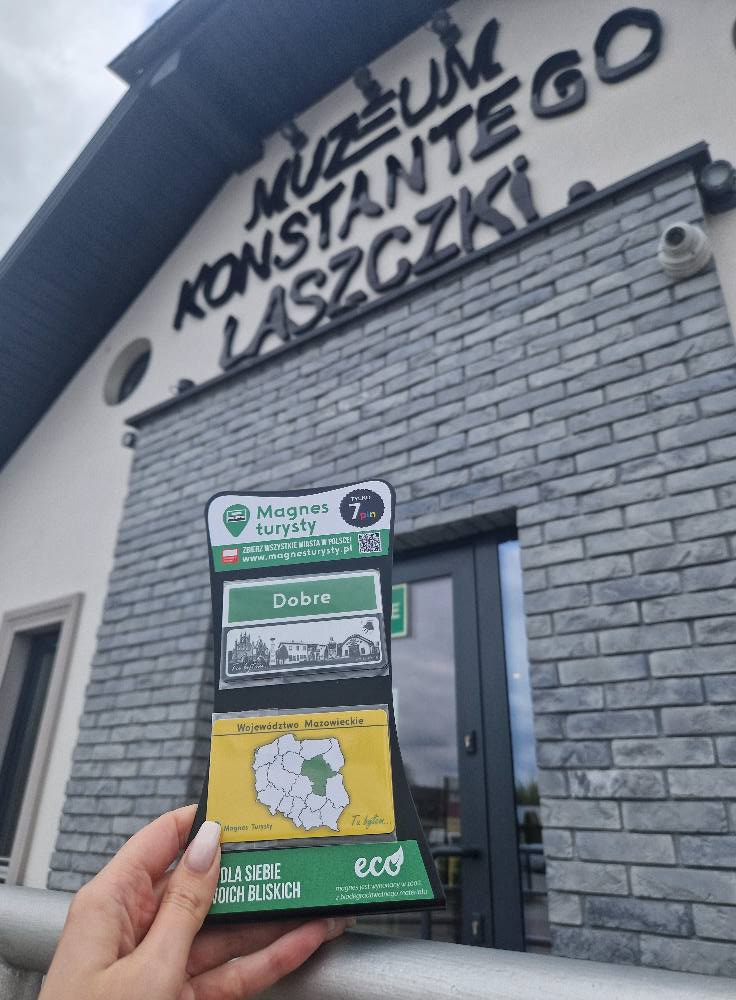 Na zdjęciu jest trzymany przez trzymany magnes przed muzeum Konstantego Laszczki. Na szarym budynku za nią dominuje napis z nazwą muzeum.