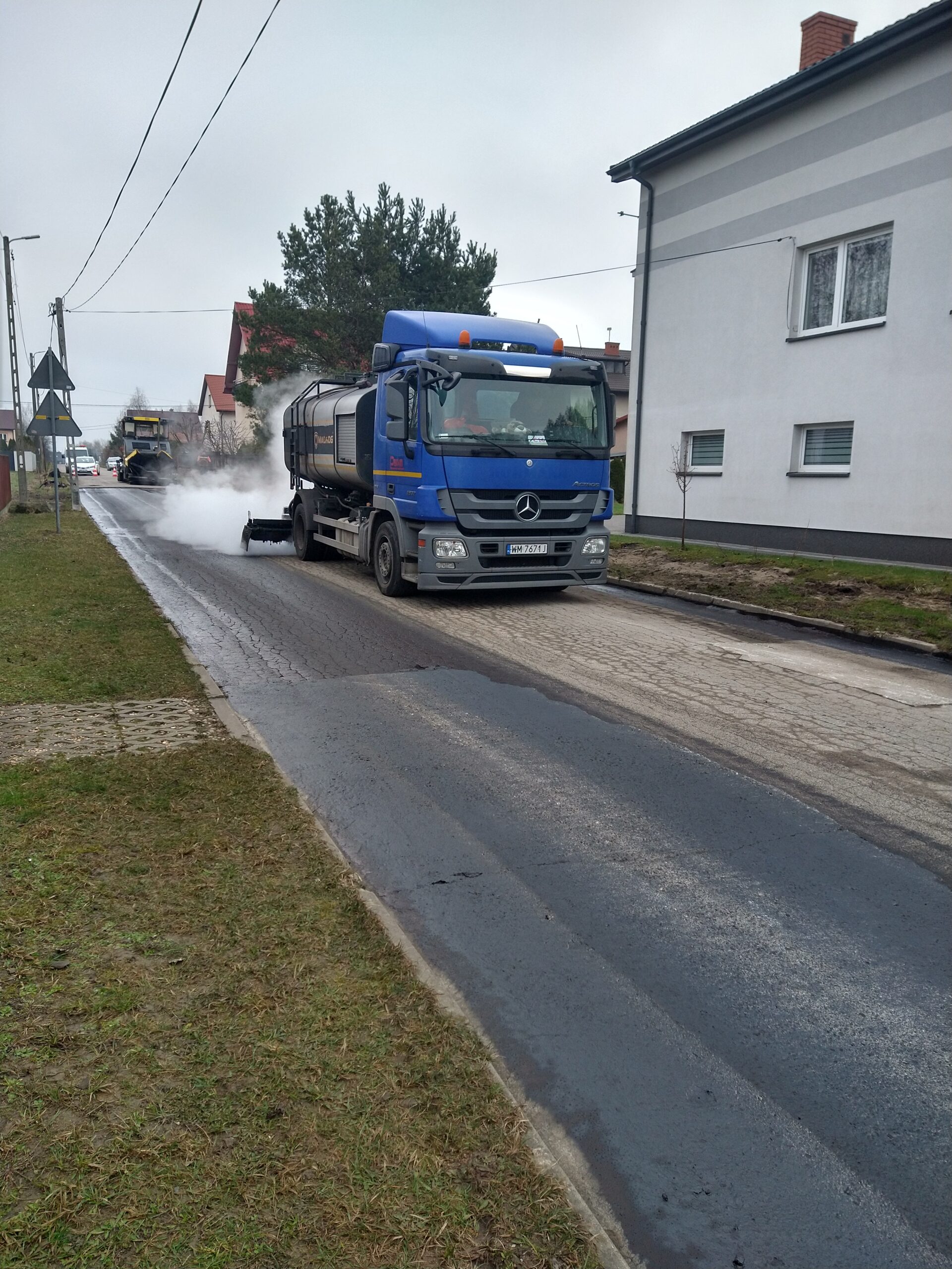 Na obraz widoczny jest samochód do czyszczenia ulicy, który rozpyla wodę na drogę. W tle można zrelaksować się domy oraz dodatkowe dodatkowe, a wszystko, co dzieje się podczas pochmurnego dnia.
