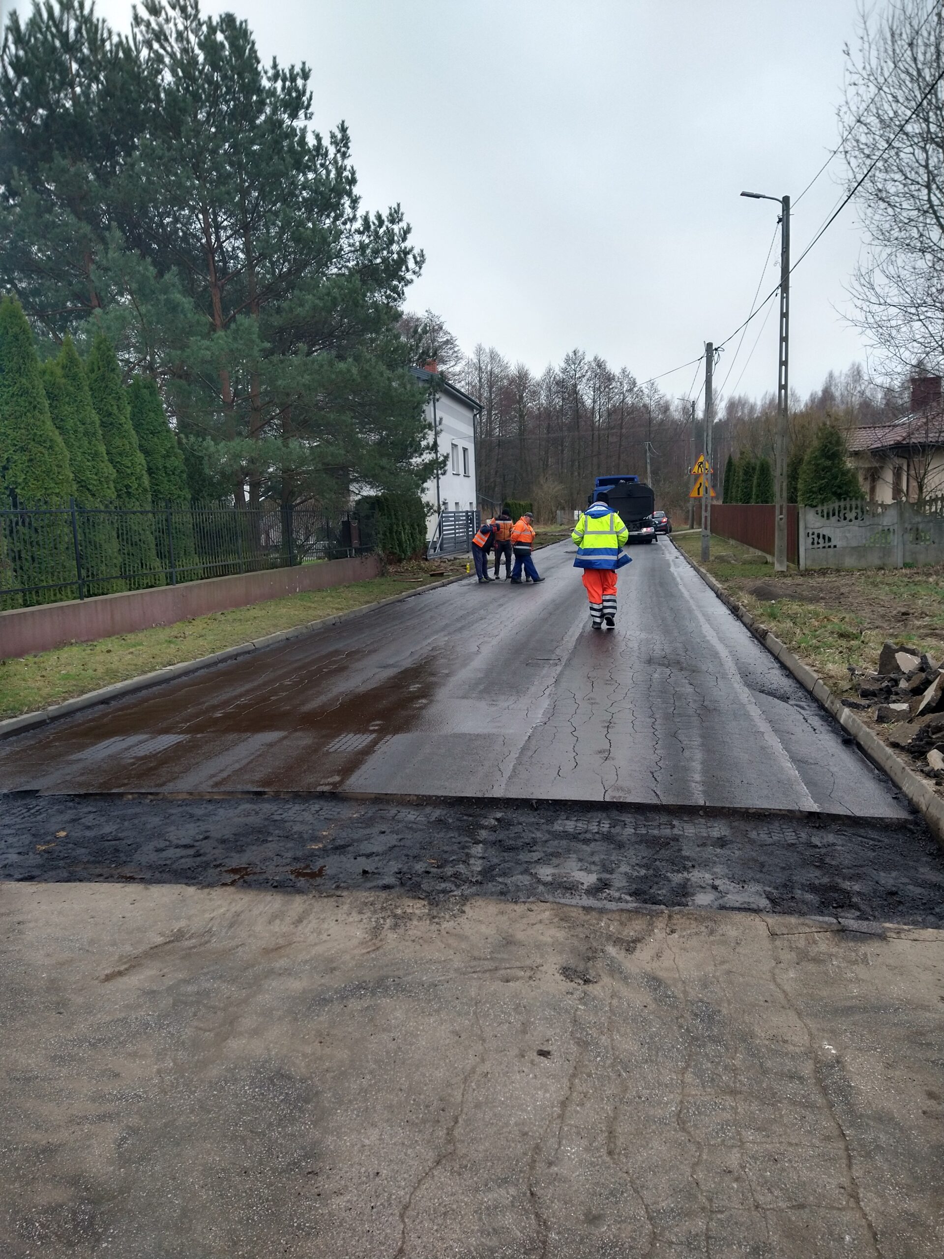 Na obrazach widać pracowników w jaskrawych kamizelkach, obsługujących maszyny na mokrej drodze podczas pracy budowlanej. W tle są widoczne, że są one mieszkalne oraz drzewa.